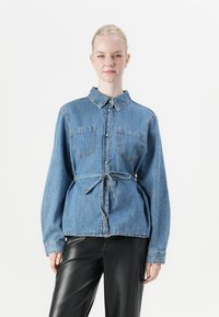 Lichtblauwe denimshirt met een klassieke kraag, twee borstzakken en een gestrikt taille. Lange mouwen met knoopsluiting en contrasterende stiknaden.
