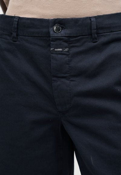 Gros plan sur la taille d’un pantalon bleu marine avec fermeture par bouton et une petite étiquette indiquant « CLOSED » sous le bouton.