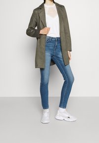 Cappotto lungo verde oliva su una maglietta in rete bianca, abbinato a jeans skinny blu e sneakers bianche. Texture lisce e linee pulite in tutto l'insieme.