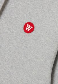 Šedá látka s jemnou textúrou, ktorá má červenú kruhovú nášivku s bielym písmenom "W". Minimalistický dizajn, bez ďalších vzorov alebo akcentov.