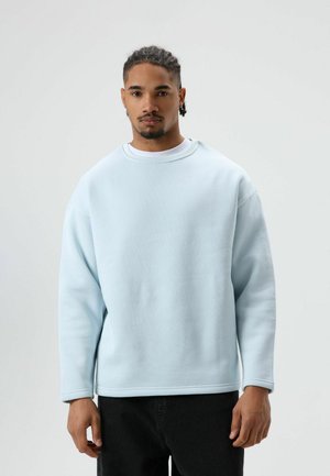 Lichtblauwe trui met een ronde hals en lange mouwen, gemaakt van zachte stof met een gladde textuur, gecombineerd met een donkere broek.