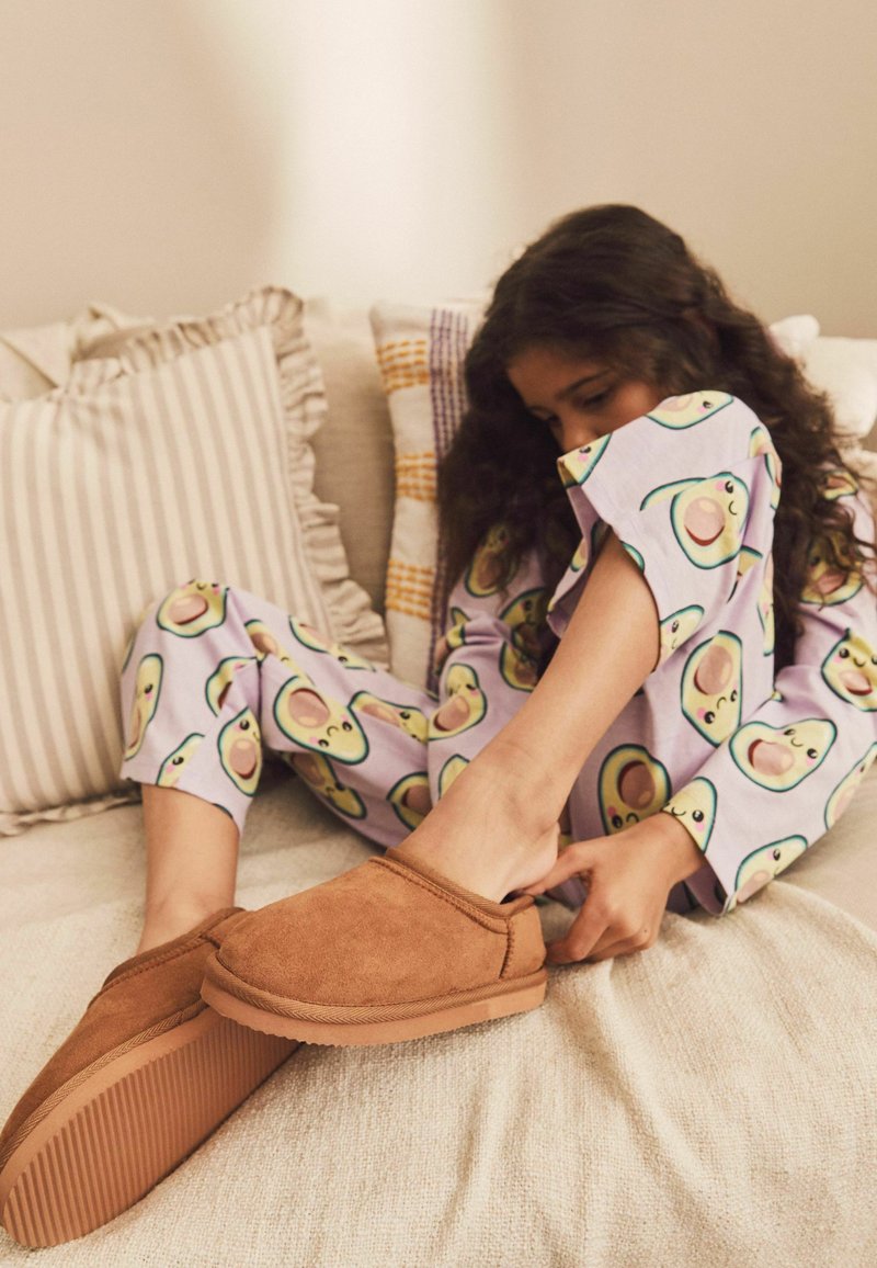 Pantuflas de ante marrón con una suela texturizada, combinadas con un pijama lila que presenta patrones de aguacates verdes y un corte relajado.