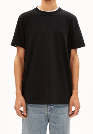 Persona con una camiseta negra lisa de manga corta y vaqueros de mezclilla azul claro, de pie frente a un fondo blanco.
