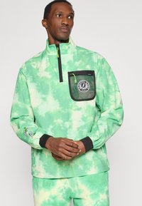 Πράσινο πουλόβερ με βαφή γραβάτας, ημί-φερμουάρ, μαύρη πορτοκαλί τσέπη από δίχτυ, ρυθμιζόμενο γιακά και ελαστικές μανσέτες. Περιλαμβάνει γραφικά σχέδια στα μανίκια.