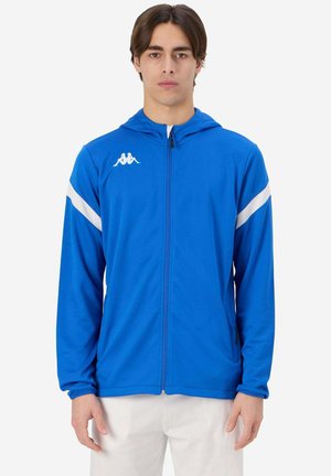 Blauwe sportjack met capuchon, gemaakt van soepele stof, met witte diagonale strepen en een klein logo op de borst. Voorzien van een rits aan de voorkant.