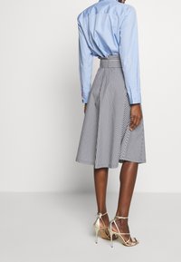 Žena oblečená v černobílé vertikálně pruhované midi sukni s páskem, světle modré košili s dlouhým rukávem a metalických stiletto sandálech, viděná zezadu.