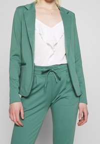 Grön stickad blazer med fällknivsklappar, parat med en vit topp med volangdetaljer och matchande gröna byxor med elastisk midja. Enkelt design.