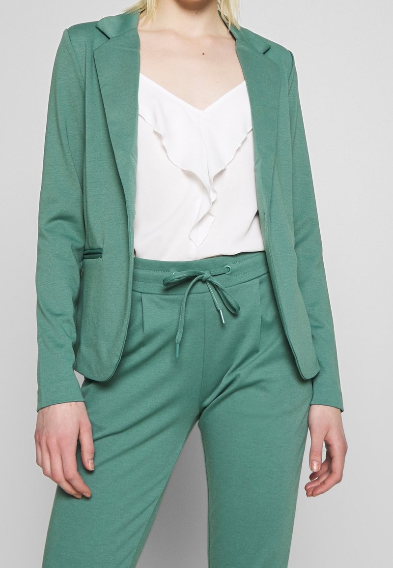 Grön stickad blazer med fällknivsklappar, parat med en vit topp med volangdetaljer och matchande gröna byxor med elastisk midja. Enkelt design.