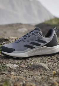 Gray and black athletic shoe with thick white sole resting on rocky outdoor terrain with blurred mountain background.