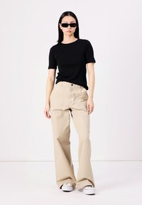 T-shirt nero a maniche corte abbinato a pantaloni beige a gamba larga, con una texture liscia. Modella che indossa occhiali da sole neri, in piedi su uno sfondo chiaro.