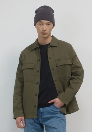 Young man wearing dark beanie, olive green buttoned jacket over black shirt, and blue jeans, standing against plain light background.
