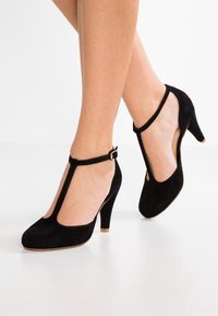 Korkokenkät, joissa on musta mokka, T-muotoiset hihnat, avoimet sivut ja pyöreä kärki. Kultasävyinen solki nilkassa.