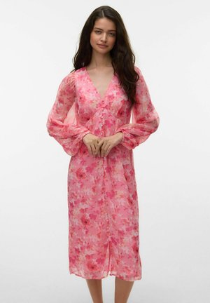 Femme portant une robe midi rose à fleurs avec de longues manches bouffantes transparentes et un décolleté en V, debout devant un fond blanc uni.