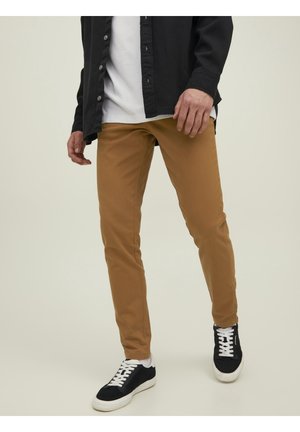 Man draagt bruine slim-fit broek, zwarte sneakers met witte veters, witte sokken, wit T-shirt en een opengeknopt zwart overhemd.