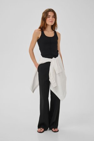 Naine seisab, kandes musta varrukateta toppi, musta laiade säärtega pükse, vöökohal seotud valget särk ja musti varbasaapaid.