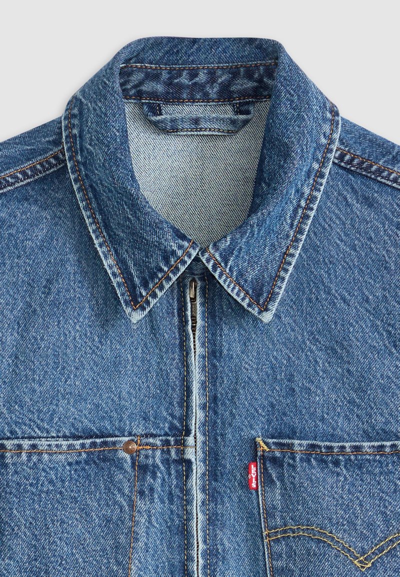 Giacca di jeans blu con collo, chiusura frontale con zip, doppie tasche sul petto e etichetta rossa Levi's. Texture distressed e cuciture a contrasto.