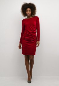 Robe en velours rouge à manches longues, avec un détail drapé à la taille et un col rond. La mannequin porte des chaussures argentées à talons hauts.