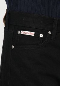 Fekete farmernadrág, piros és fehér "Calvin Klein" felirattal, ezüst színű kiegészítőkkel, sima felülettel és diszkrét varrási részletekkel.