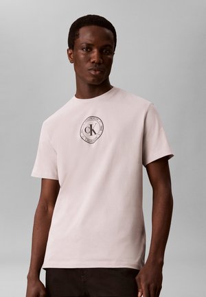 Νέος άνδρας φοράει ένα ανοιχτό μπεζ T-shirt Calvin Klein με κυκλικό λογότυπο στο στήθος, στέκεται μπροστά σε απλό γκρι φόντο.