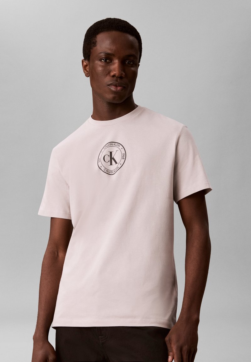 Νέος άνδρας φοράει ένα ανοιχτό μπεζ T-shirt Calvin Klein με κυκλικό λογότυπο στο στήθος, στέκεται μπροστά σε απλό γκρι φόντο.