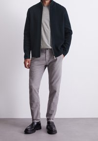 Giacca verde scuro con zip, maglietta grigia a girocollo, pantaloni grigi chiari e mocassini neri. Tessuti lisci e design semplice.