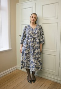 Modrá kvetinová šaty s výstrihom do V, trojštvrťovými rukávmi a voľným strihom, skombinované s čiernymi vysokými podpätkami.