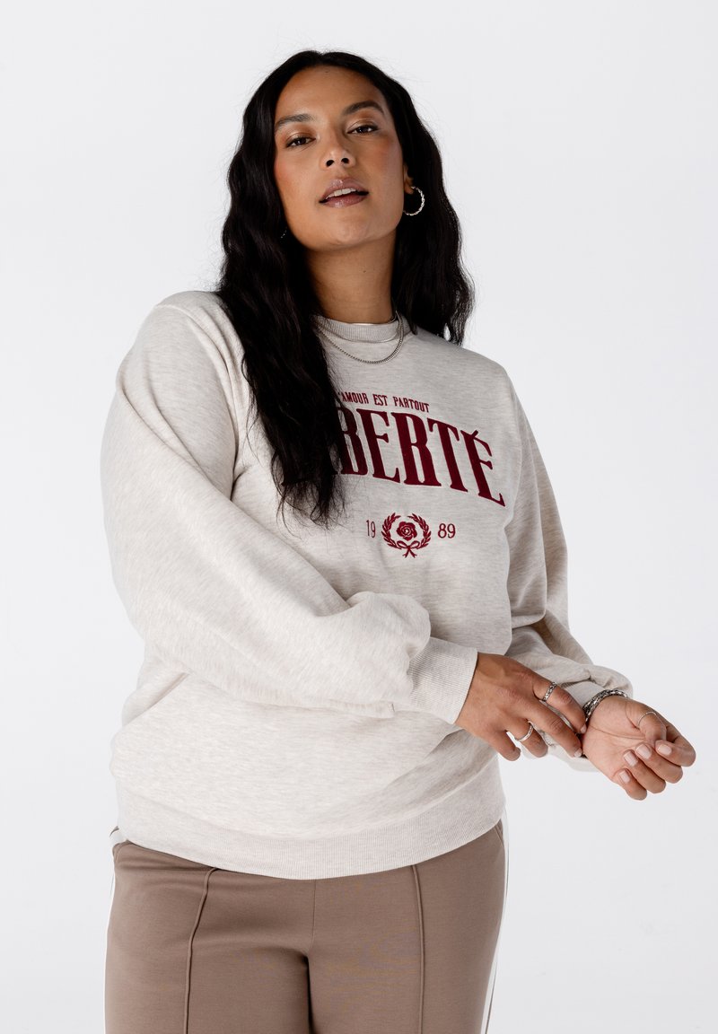 Femme avec de longs cheveux noirs portant un sweat-beige avec le texte rouge "LIBERTÉ" et un pantalon marron, ajustant sa manche sur un fond blanc.
