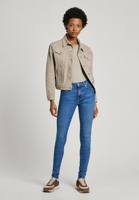 Chaqueta de mezclilla beige con dos bolsillos frontales, combinada con una blusa verde clara y jeans ajustados azul oscuro. Zapatillas marrones y crema completan el atuendo.