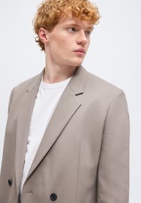 Νέος άνδρας με κατσαρά κόκκινα μαλλιά που κοιτάζει προς τα δεξιά, φοράει ένα ανοιχτό καφέ σακάκι πάνω από ένα λευκό t-shirt με στρογγυλή λαιμόκοψη, μπροστά σε απλό φόντο.