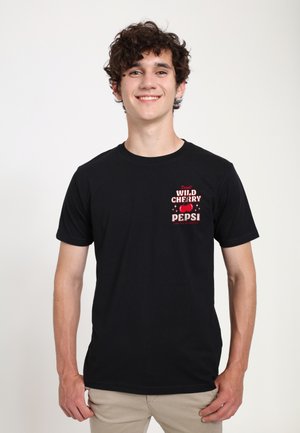 Jeune homme aux cheveux bouclés portant un T-shirt noir avec l'inscription rouge "Wild Cherry Pepsi" et des images de cerises, debout sur un fond blanc.