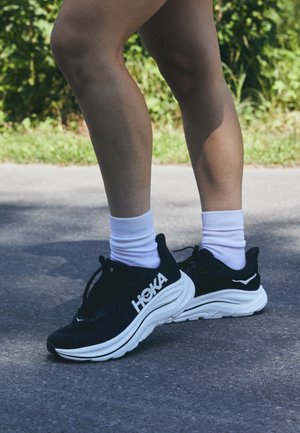 Black running shoes with white soles, featuring a prominent white logo and laces. Worn with white crew socks on a paved surface.