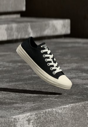 Μαύρο αθλητικό παπούτσι χαμηλού τύπου με λευκά κορδόνια και σόλα, αιωρούμενο πάνω από γκρίζα σκυρόδεμα και σκάλες στο φόντο.