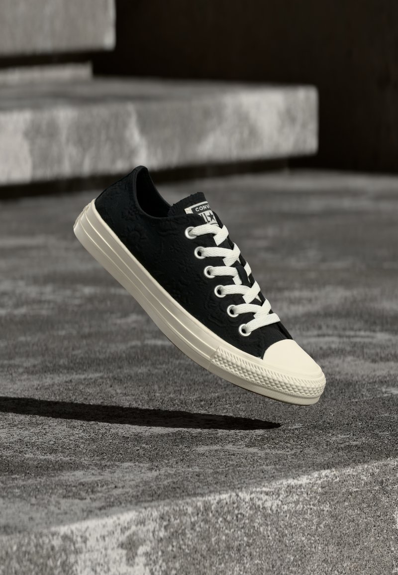 Μαύρο αθλητικό παπούτσι χαμηλού τύπου με λευκά κορδόνια και σόλα, αιωρούμενο πάνω από γκρίζα σκυρόδεμα και σκάλες στο φόντο.