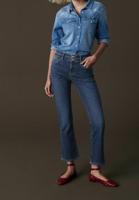 Denim-paita, jossa on kaksi rintataskua ja brodeerauksia, yhdistettynä korkeavyötäröisiin sinisiin farkkuihin ja punaisiin ballerinoihin, joissa on valkoisia pilkkuja.