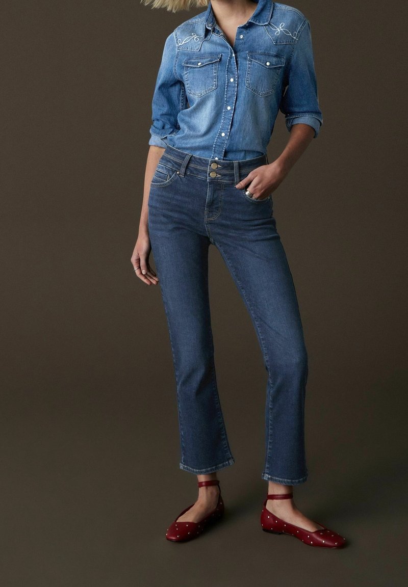 Denim-paita, jossa on kaksi rintataskua ja brodeerauksia, yhdistettynä korkeavyötäröisiin sinisiin farkkuihin ja punaisiin ballerinoihin, joissa on valkoisia pilkkuja.