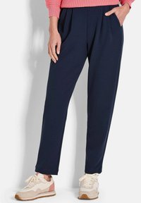 Femme portant un pantalon plissé bleu marine et des baskets blanches avec des accents roses, se tenant avec une main dans la poche et l'autre main détendue.