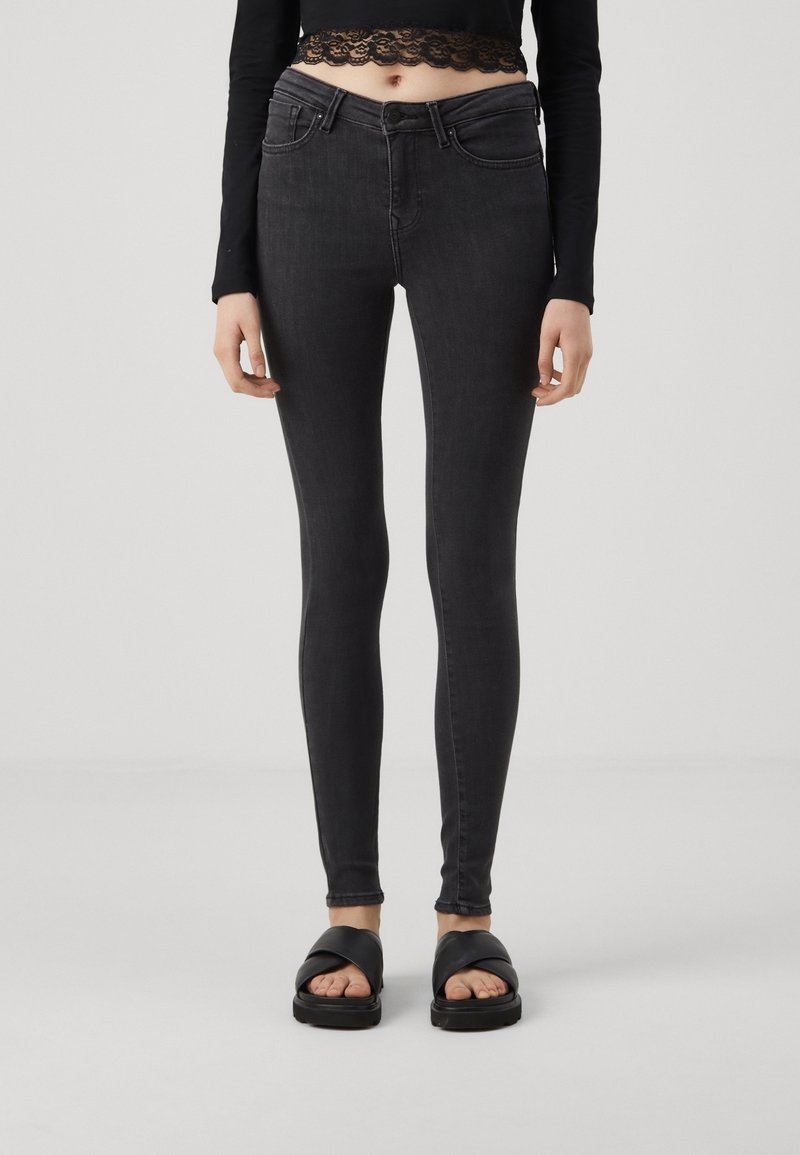 Jeans negros ajustados con una textura suave, que cuentan con un diseño clásico de cinco bolsillos y una cintura de altura media, combinados con sandalias negras tipo slider.