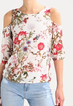 Blouse blanche épaules dénudées avec imprimé floral rose, rouge et violet, portée avec un jean bleu clair déchiré.