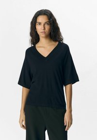 Black v-neck sweater, short sleeves, knit texture, relaxed fit. Paired with black trousers for a monochromatic look. Simple design.