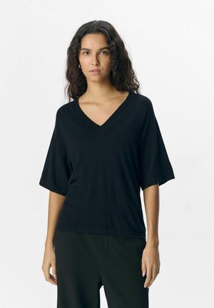 Suéter negro de cuello en V, de manga corta, con textura de punto y corte relajado. Combinado con pantalones negros para un look monocromático. Diseño simple.