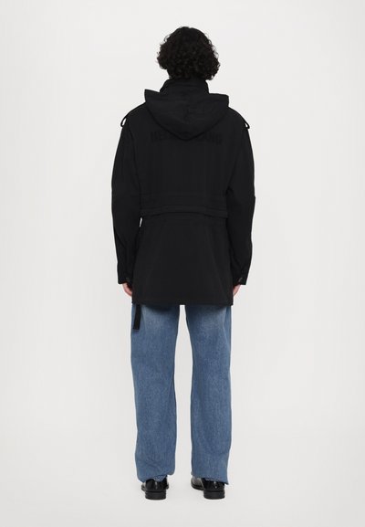 Zwarte gevoerde jas met een ceintuurlint in de taille, met een gladde textuur en een matte afwerking. Gecombineerd met lichtblauwe denimbroek.