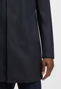 Cappotto lungo blu navy in tessuto testurizzato, con una tasca laterale e dettagli delle maniche lisci. Vestibilità sartoriale con orlo dritto.