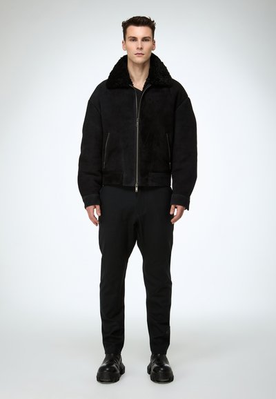 Veste en daim noire avec un grand col en fourrure noire douce, dotée d'une fermeture éclair frontale et de deux poches latérales. Portée avec un pantalon noir et des bottes épaisses.