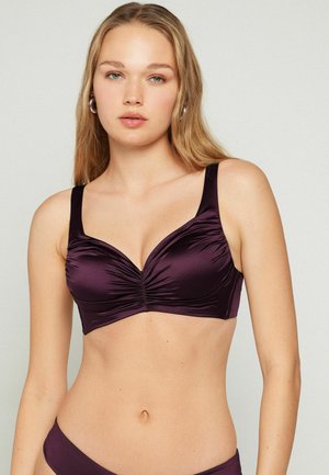 Young woman with long blonde hair wearing a dark purple ruched bra and matching underwear, standing against a plain light background.
