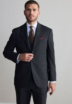 Sötétszürke öltönyös férfi mintás nyakkendővel igazítja zakóját egyszínű világos háttér előtt.