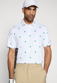 Λευκό πόλο μπλουζάκι με μικρά πράσινα λογότυπα, διαθέτει γιακά και κοντά μανίκια. Φορεμένο με καφέ παντελόνι και κρατώντας μια ρακέτα γκολφ.