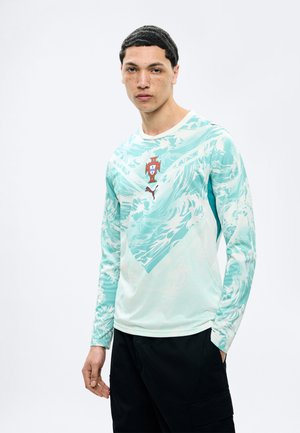 Νεαρός άνδρας που φοράει πουκάμισο με μακριά μανίκια σε ανοιχτό μπλε και λευκό με το σήμα της πορτογαλικής ποδοσφαιρικής ομάδας και λογότυπο Puma, στέκεται με το ένα χέρι στην τσέπη.