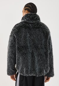 Eine Person mit geflochtenem Haar trägt eine dicke schwarz-weiße, gefleckte Kunstfelljacke, von hinten gesehen, kombiniert mit schwarzen Hosen mit weißen Streifen.