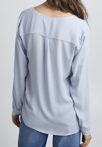 Blusa azzurra a maniche lunghe con scollatura arrotondata, texture seta, e orlo curvo. Presenta una vestibilità leggermente morbida e polsini con bottoni.