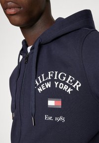 Haut de survêtement zippé marine avec le texte et le logo blanc "HILFIGER NEW YORK". Comprend des cordons de serrage, des fixations en métal argenté et un accent graphique de l'année d'établissement 1985.